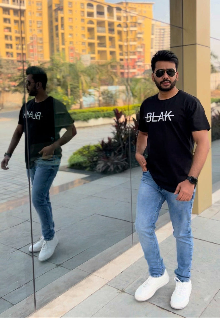 Man wearing a black t-shirt with 'BLAK' text, standing in front of a reflective surface with buildings in the background.